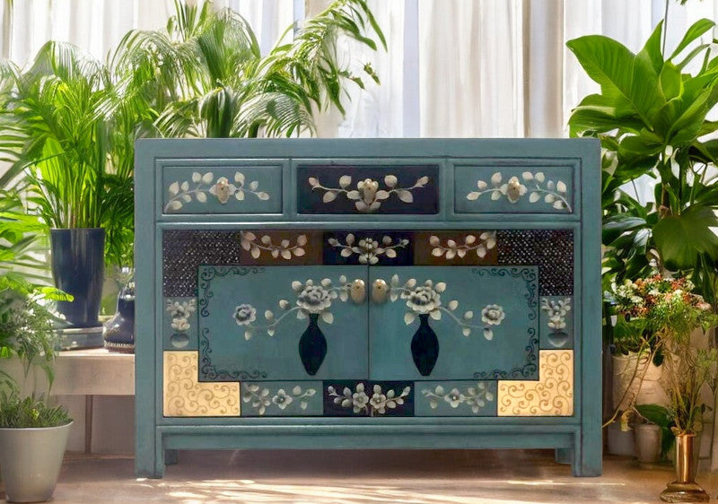 Kommode im floralen Design mit drei Schubladen und Doppeltüren, umgeben von üppiger Pflanzenkulisse vor hellen Vorhängen.