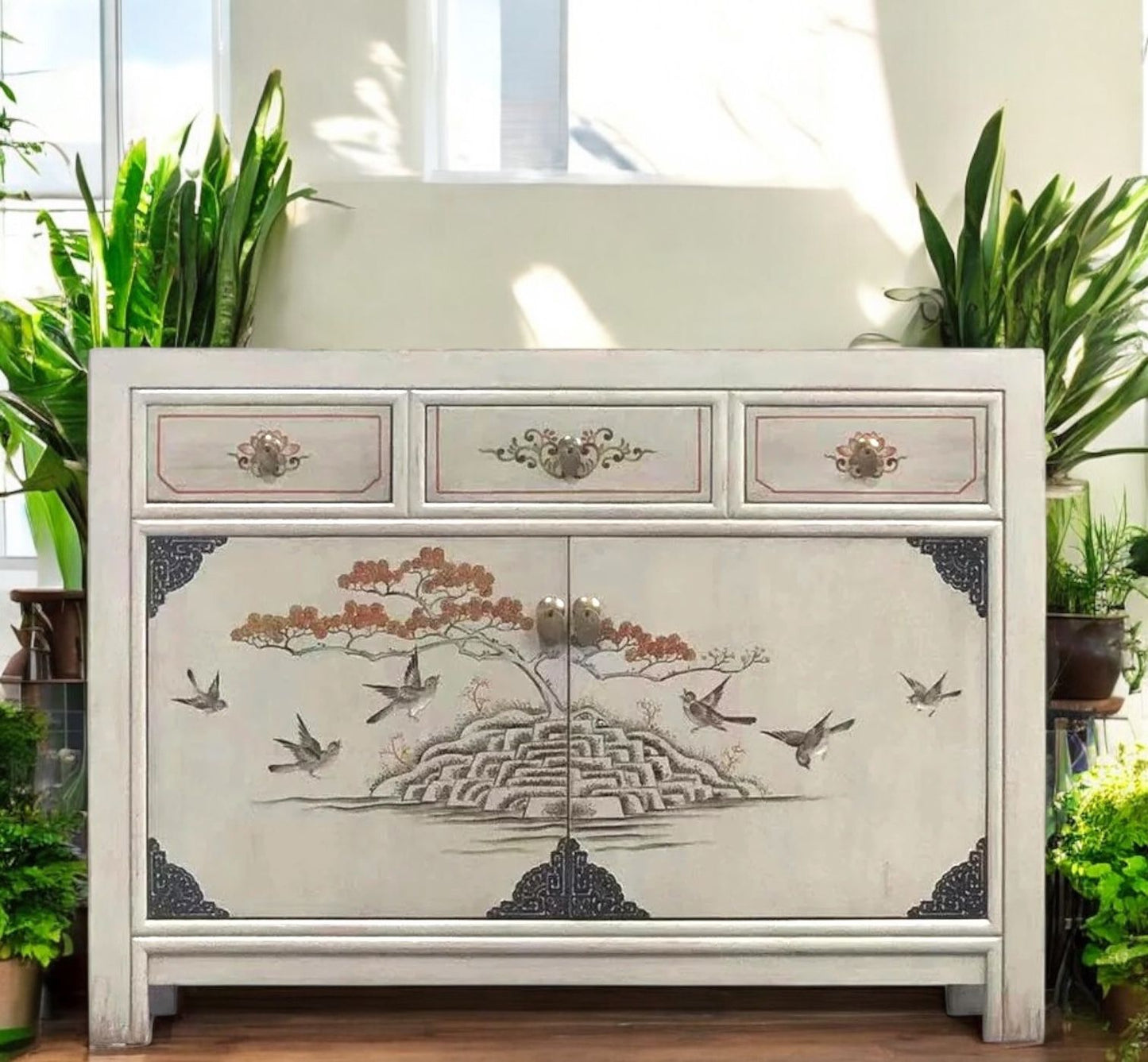 Vintage-Kommode im hellen Raum zwischen Pflanzen, mit asiatischem Vogel- und Baum-Motiv auf der Türfront, ideal inszeniert im Sonnenlicht.