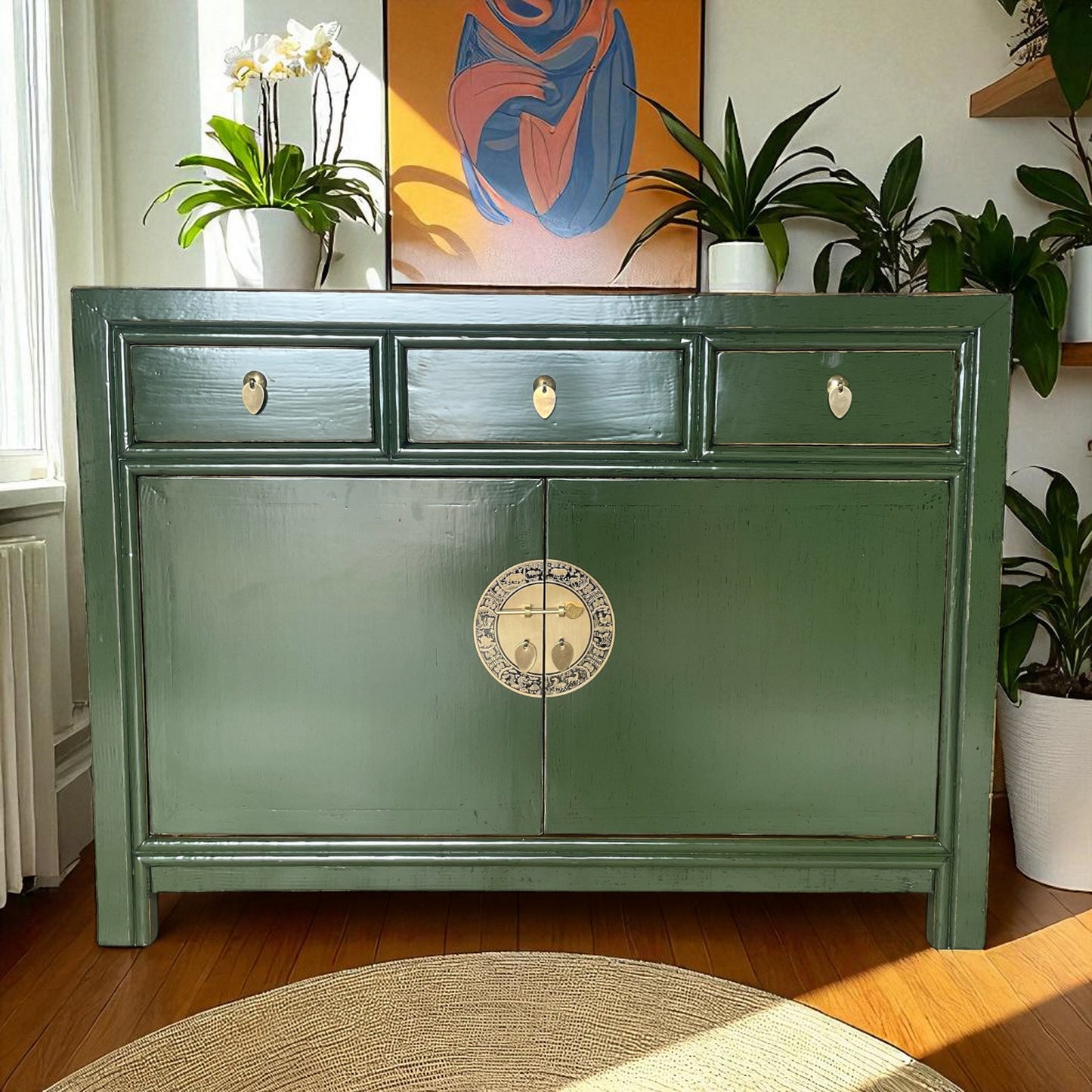 Elegante grüne Kommode vor einem Fenster, umgeben von Pflanzen und moderner Kunst, in sonnendurchflutetem Raum.