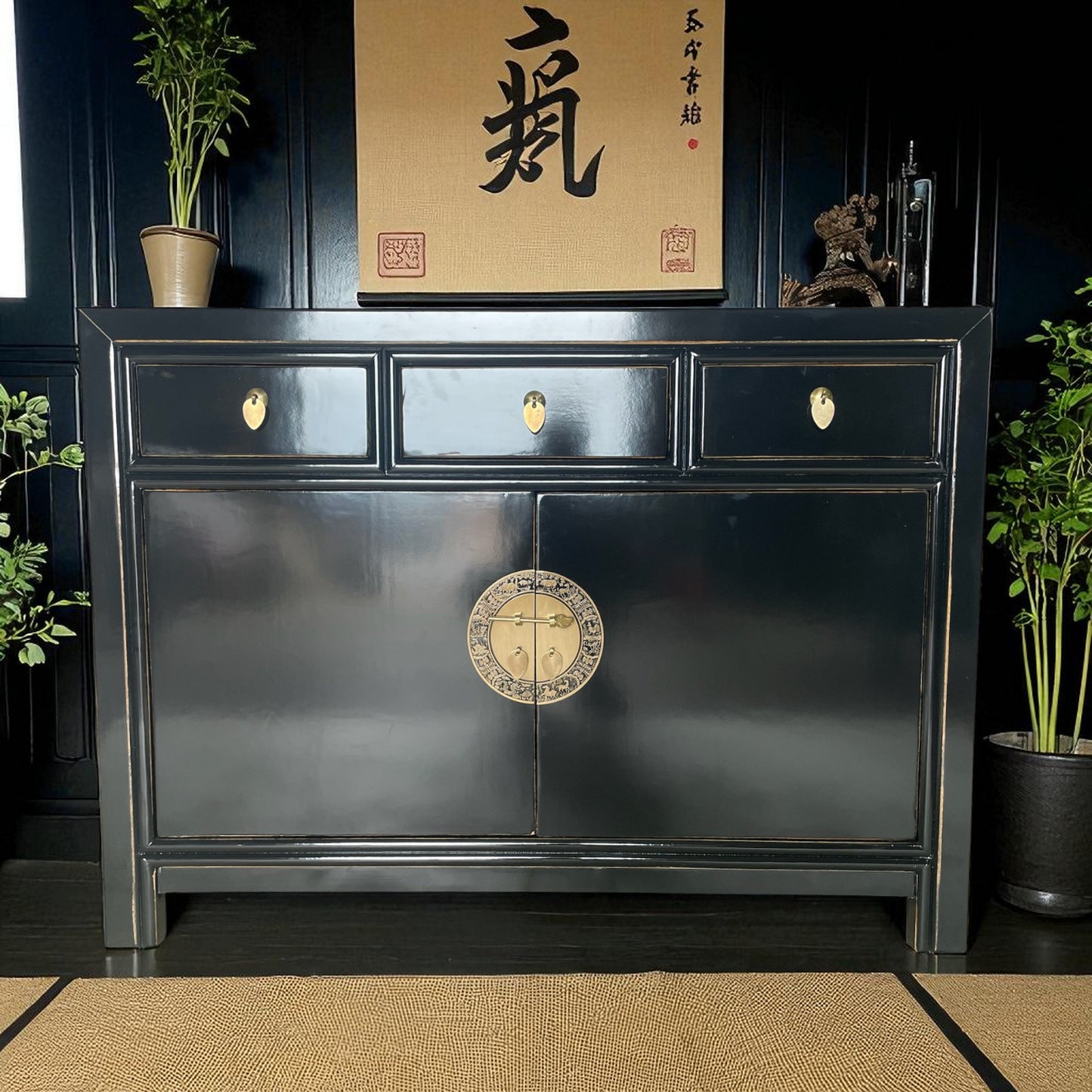 Schwarze asiatische Kommode mit drei Schubladen und vergoldetem Rundbeschlag, stilvoll inszeniert vor schwarzer Wand mit Deko und Pflanzen.