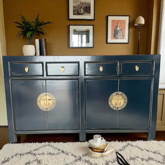 Dunkelblaues Sideboard mit goldenen Rundbeschlägen, platziert in einem warm eingerichteten Wohnzimmer mit Teppich und Wandbildern.