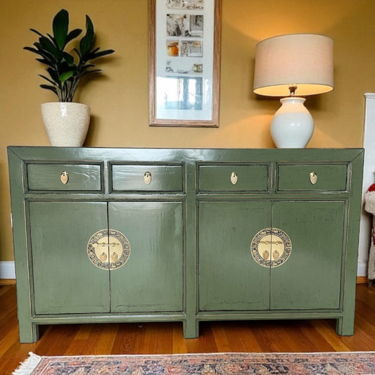 Grünes Sideboard mit goldfarbenen runden Beschlägen in einem Wohnzimmer mit Pflanze, Tischlampe und Wanddeko.