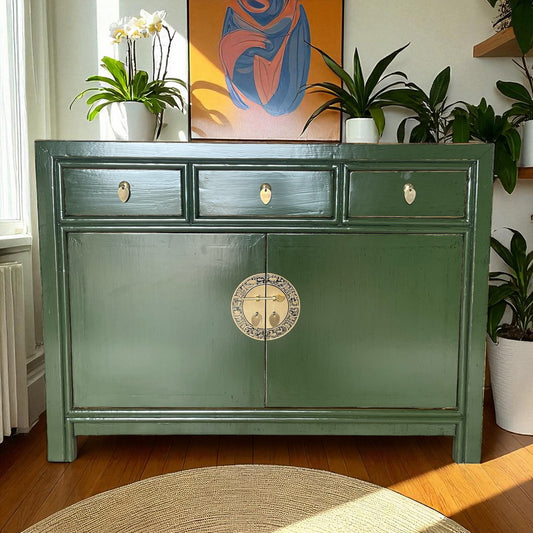 Elegante grüne Kommode vor einem Fenster, umgeben von Pflanzen und moderner Kunst, in sonnendurchflutetem Raum.