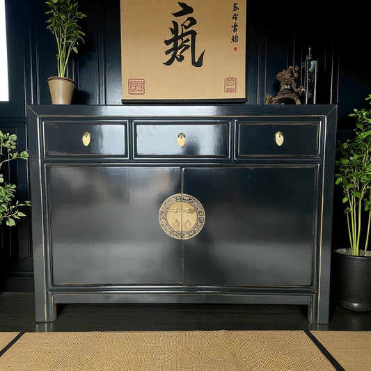 Schwarze asiatische Kommode mit drei Schubladen und vergoldetem Rundbeschlag, stilvoll inszeniert vor schwarzer Wand mit Deko und Pflanzen.